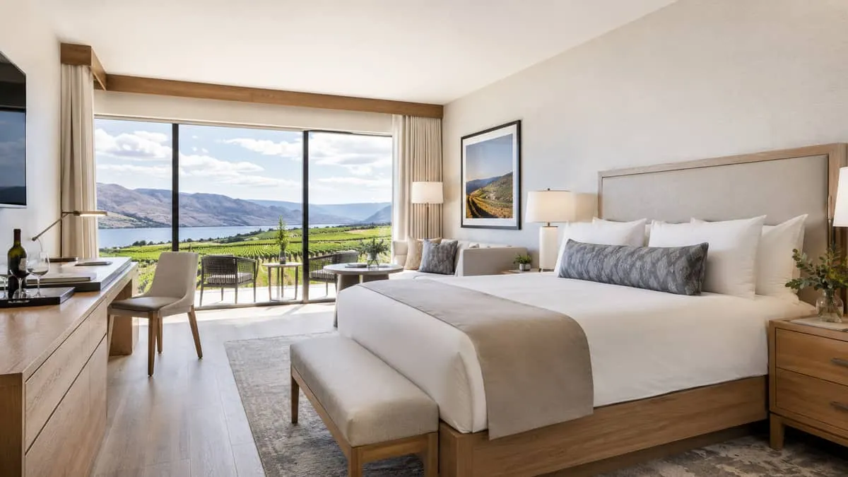 an Okanagan-style hotel room or suite with natural wood furniture, warm linen upholstery, and a window view of vineyard 