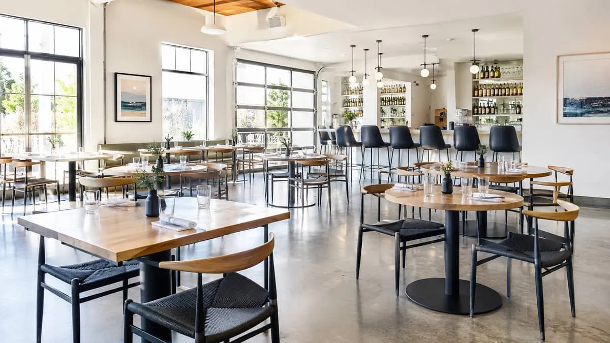 a mix of round and square restaurant tables with commercial table bases on a polished concrete floor, barstools visible 
