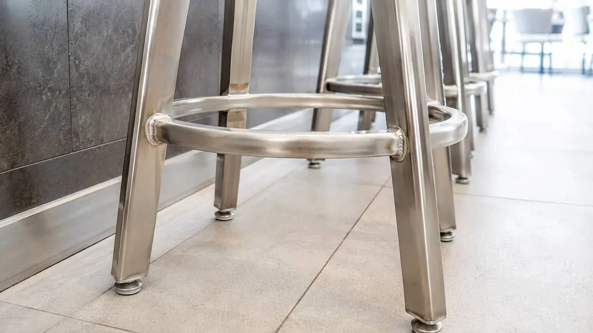 close-up of a commercial barstool footrest and base showing weld quality and floor glide hardware, with a tile floor typ