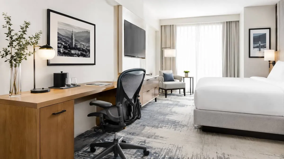 a boutique hotel guest room desk area with a solid wood writing desk, task chair with proper lumbar support, and a bedsi