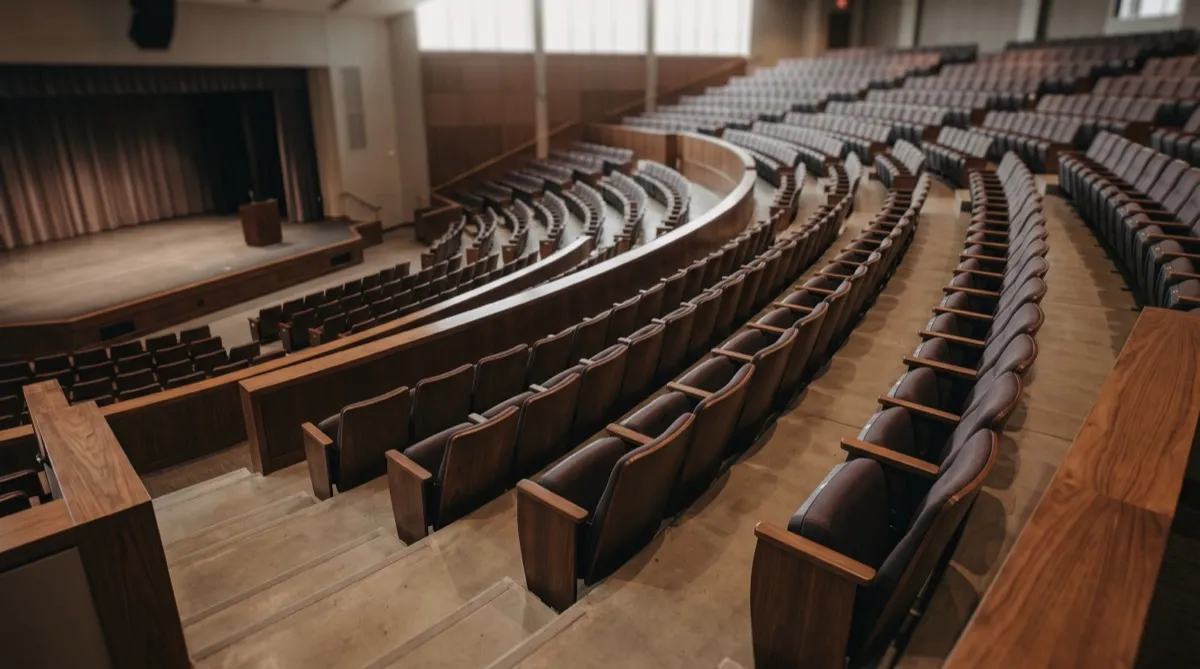a raked auditorium tier viewed from a middle row looking down toward a distant stage platform, the sloped floor and stepped rows emphasizing